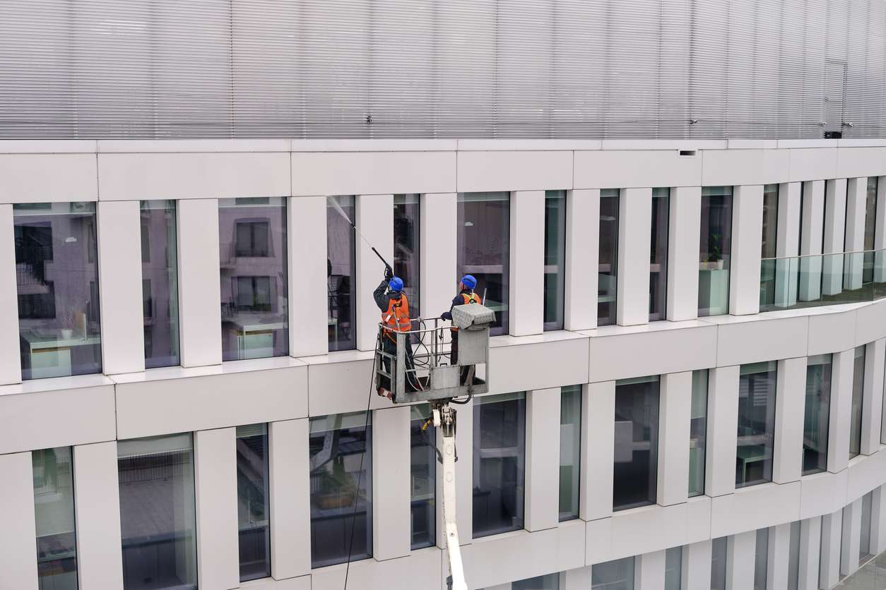 Two workers wearing safety harness wash office building facade at height standing in a crane cradle or aerial platform using pressure washer and mops